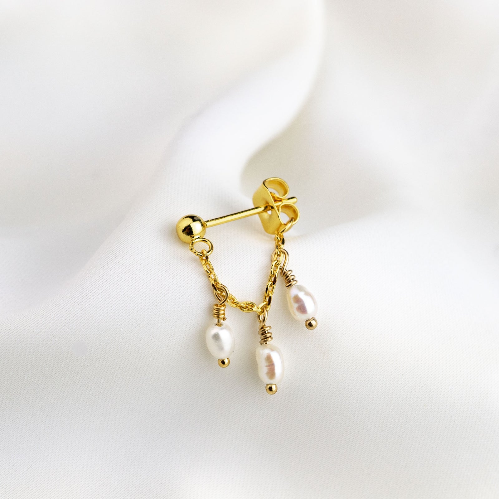 A single gold-plated earpin with a chain and pearl embellishments on a white fabric background.